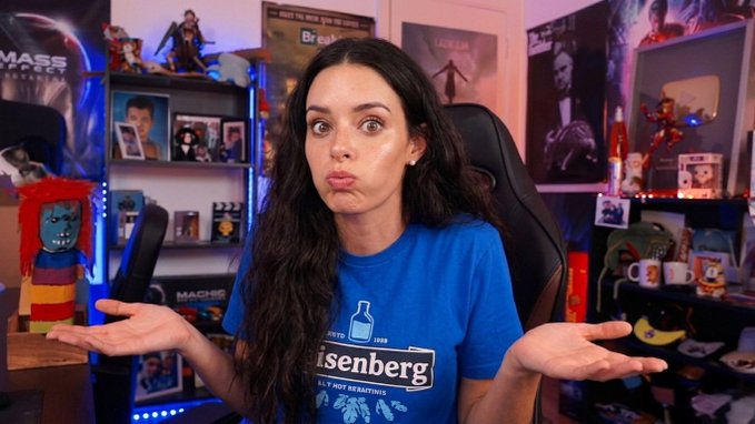 A woman in a blue "Heisenberg" t-shirt makes a confused, shrugging face with her palms up, surrounded by pop culture collectibles including Mass Effect and Breaking Bad memorabilia. Her "I don't know" expression and Heisenberg shirt (a Breaking Bad reference) visually capture the caption's sentiment of being exasperated and indifferent to the noise of crypto Twitter's endless commentary.