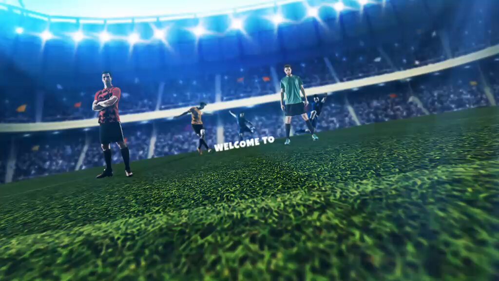 The image features a soccer field with several players standing around. The main focus of the image is a group of people posing for a photo near the center of the field. There are at least six visible individuals, including two men wearing red shirts and one man in a green shirt. They appear to be enjoying their time together on the field.

The background of the image shows an empty stadium with bright lights illuminating the area. The players seem to be standing near the center of the field, possibly waiting for their turn or taking a break from playing.