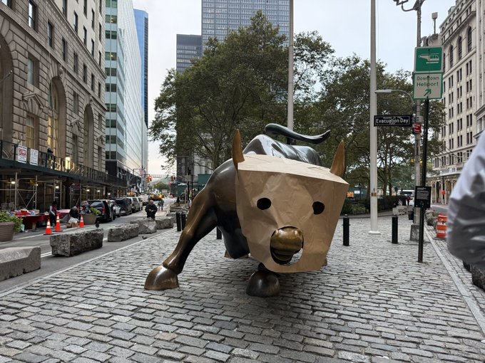 The bronze Charging Bull statue on Broadway has been humorously adorned with a brown paper bag over its head, with a "Election Day" street sign visible nearby, capturing a scene that's been interpreted as a viral meme moment tied to the WIF crypto token's cultural commentary.