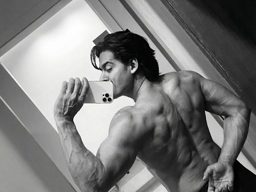 A black-and-white photo shows a muscular, shirtless man taking a mirror selfie with a smartphone.