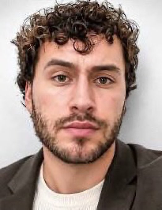 A close-up portrait of a young man with curly brown hair, a short beard, and brown eyes, wearing a dark jacket over a white shirt against a light background.