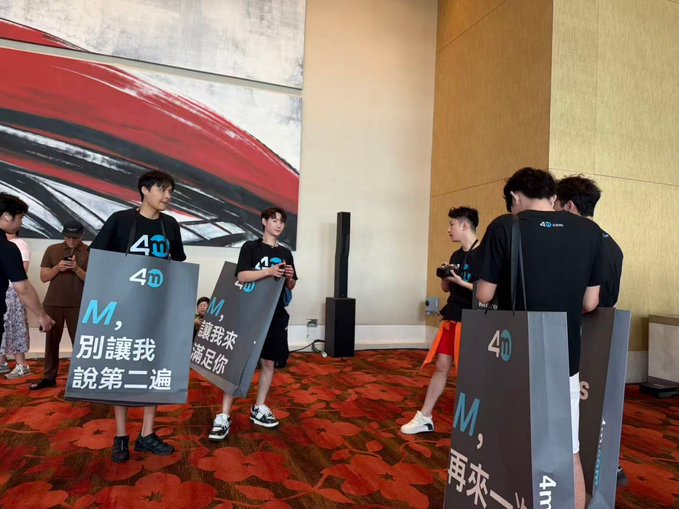 A group of people in matching black t-shirts hold large signs reading '4D' and 'M, 別讓我說第二遍' (M, don't make me say it twice) at what appears to be an indoor event.