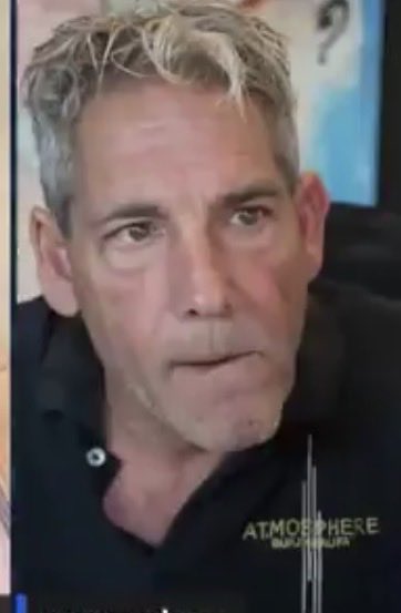 A close-up photo of a middle-aged man with messy grey hair wearing a dark shirt, appearing to speak, with the watermark 'ATMOSPHERE' visible at the bottom right.