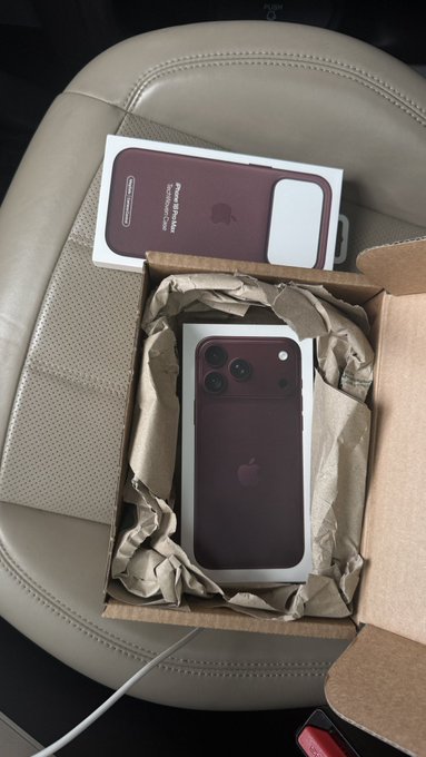 An iPhone 17 Pro or iPhone 17 Pro Max in a deep reddish-purple color sits in its packaging inside a cardboard box on a car seat, with the lid showing the text "iPhone Pro Max" and "Designed by Apple in California."