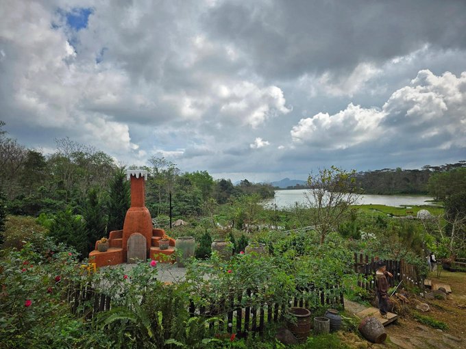 A scenic landscape view of a lush garden with a unique reddish-brown brick oven structure in the foreground, looking out over a lake and forested hills under a cloudy sky.