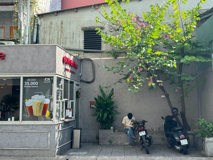 A street corner scene features a man looking at his phone while seated on a small wall next to a parked motorcycle, with a coffee shop advertising a '35,000 Holiday Season' promotion visible on the left.