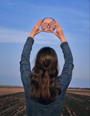 A woman viewed from behind holding up a basketball, wearing a gray long-sleeve shirt, standing in a field under a clear blue sky.