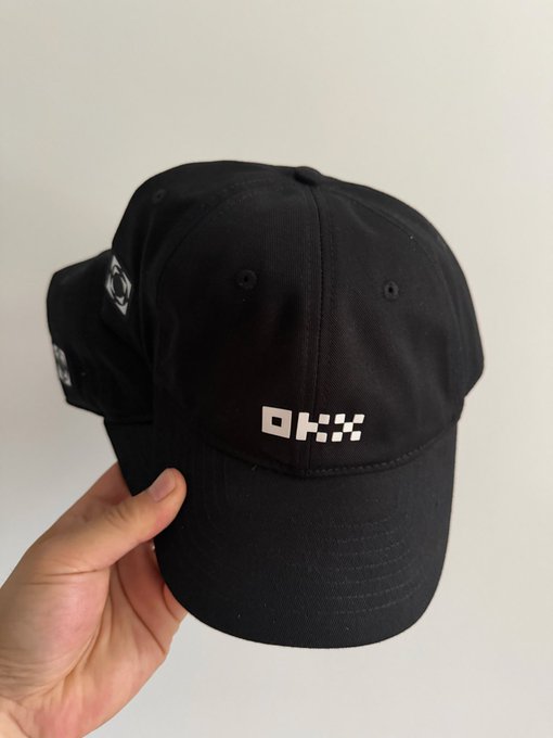 A hand holding a black baseball cap with a white pixelated 'OKX' logo embroidered on the front, with a second similar cap visible in the background.