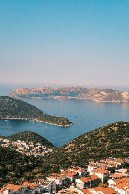 A scenic coastal view featuring a bay with a forested peninsula, a small village with white buildings and red tile roofs, and mountains in the hazy distance under a clear blue sky.