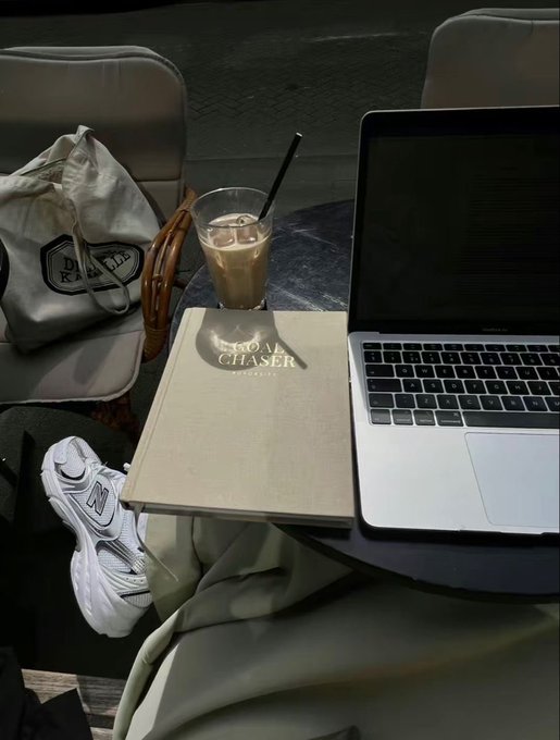 A cozy nighttime scene at an outdoor café with a MacBook Pro laptop, an iced coffee, a Royal Chaser menu, a white sneaker resting up, and a branded drawstring bag on an adjacent chair.