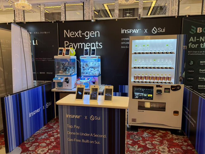 The image shows the INSPAY x Sui "Next-gen Payments" booth at the WebX2026 conference, featuring a display counter with two miniature UFO catcher (claw) machines and a vending machine, along with smartphones for tap-to-pay demos. The booth promotes gas-free Sui stablecoin payments with the tagline "Tap, Pay. Done In Under A Second," allowing attendees to use Apple ID or Google accounts to experience the futuristic payment system firsthand.
