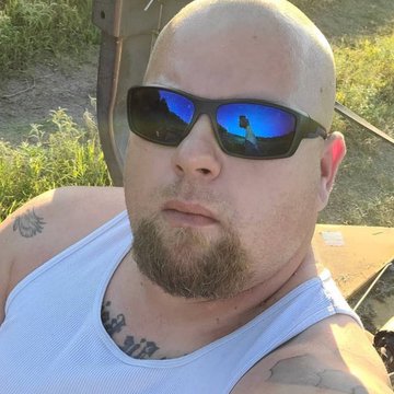 A bald man with a goatee, wearing dark sunglasses reflecting the sky, and a light blue tank top, is taking a selfie outdoors with a piece of machinery visible in the background. He has tattoos on his shoulder and chest, including a visible spider design.