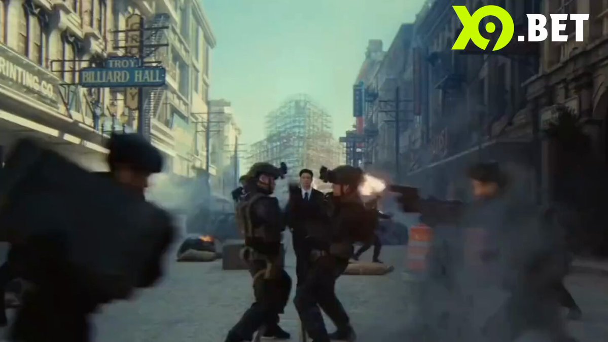 The image shows a group of people dressed in military uniforms standing in front of buildings, possibly engaged in combat or an intense situation. The scene appears to be set in a city environment with various elements such as cars and traffic lights visible around them. There is also text that reads "X9.bet" and "X.9.bet", which suggests the image might be related to a video game or promotional material for a gaming platform.