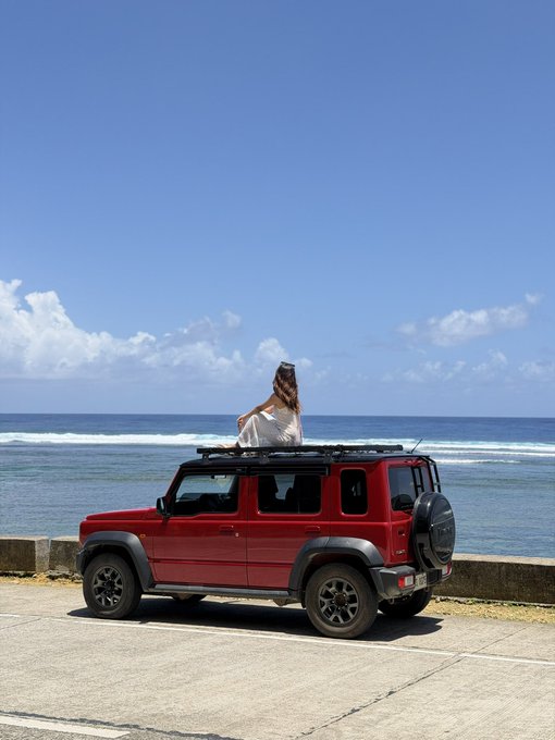 A woman with sunglasses perched on her head sits atop a red Suzuki Jimny SUV, gazing out at the ocean under a bright blue sky. The image evokes the "new beginnings" mentioned in the caption, with the open road and sea symbolizing the exciting, busy month ahead leading up to CoinfestAsia.