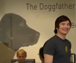 A photo of a smiling young man in a gray t-shirt standing in front of a wall featuring a graphic of a dog and the text 'The Dogfather'.