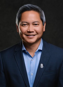 A professional headshot of a smiling middle-aged Asian man with dark hair, wearing a navy blazer over a light blue button-up shirt with a small lapel pin, set against a dark backdrop—likely Phong Le, the CEO of Strategy (formerly MicroStrategy/$MSTR), pictured in connection with his recent interview addressing questions about the company's $STRC dividend.