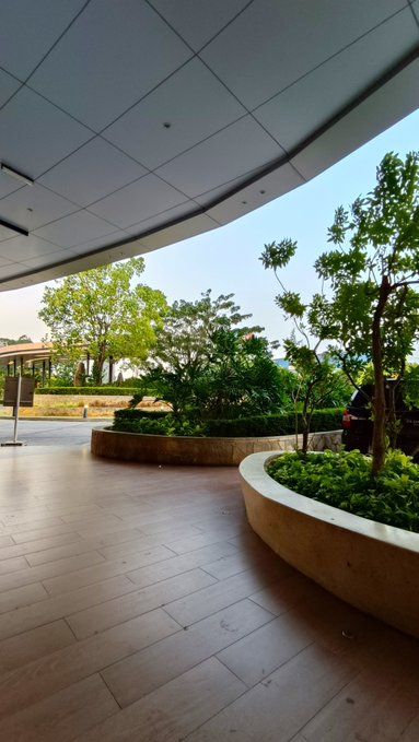 A photo looking outward from beneath a curved building overhang onto a tiled patio with landscaped greenery and trees.