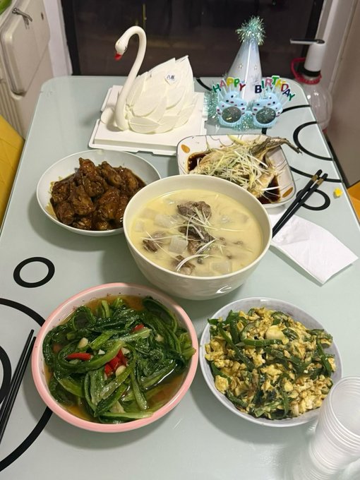 A festive birthday meal spread on a glass table featuring a white swan-shaped cake, a 'HAPPY BIRTHDAY' party hat, and dishes including braised meat, soup with noodles, steamed fish with ginger, sautéed greens, and a scramble of eggs with vegetables.