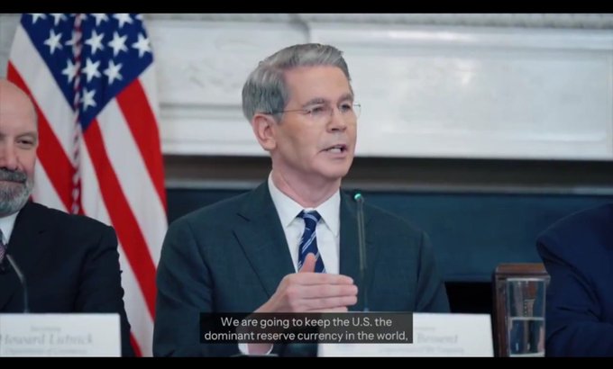 A man in a suit and tie speaks at a podium with an American flag behind him, with subtitles reading 'We are going to keep the U.S. the dominant reserve currency in the world.'
