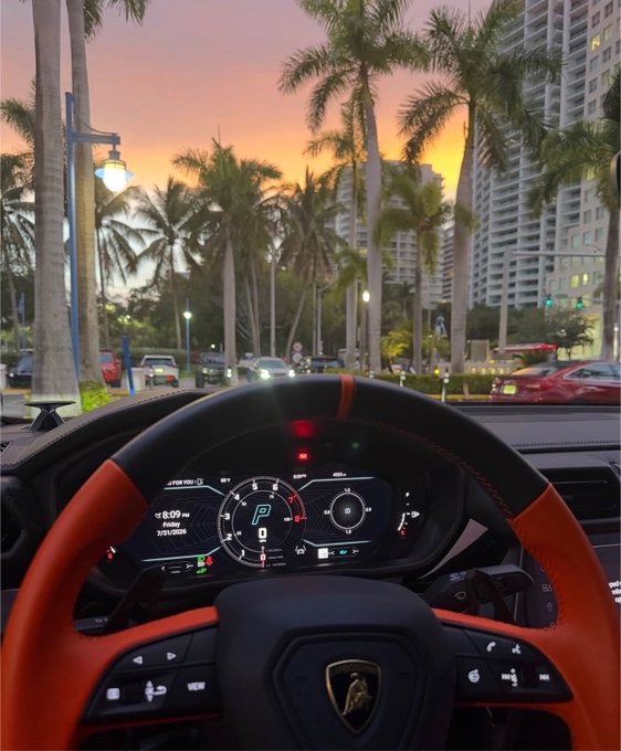 A first-person view from inside a Lamborghini sports car, showing the digital dashboard in park (P) and a sunset view of a palm-tree-lined street with buildings and traffic.