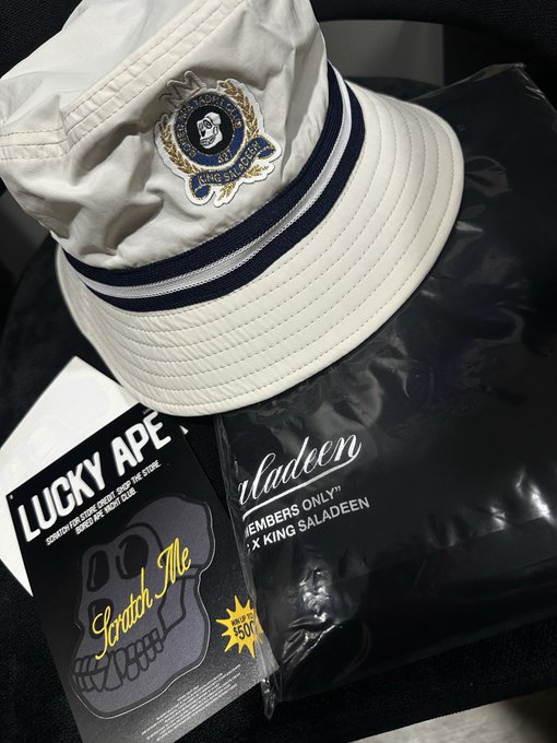 Streetwear merchandise including a cream bucket hat with a King Saladeen crest patch, alongside 'Lucky Ape' Scratch Me and 'Members Only' King Saladeen branded packaging.