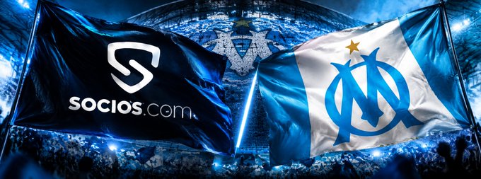 Two flags waving against a stadium background: one black flag with the "Socios" logo and another white flag with the Olympique de Marseille football club crest.
