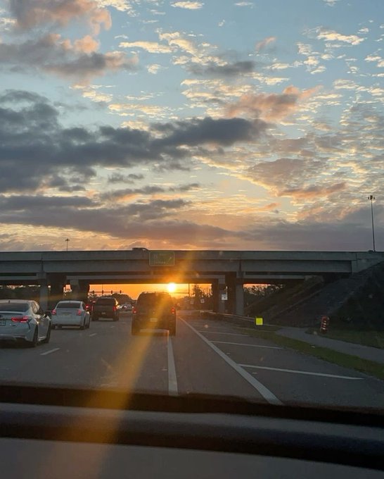 A sunset view from inside a vehicle shows cars on a highway passing under an overpass, with the sun low on the horizon.
