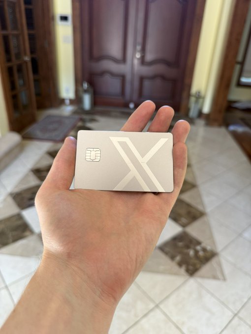 A hand holding a metallic silver card with an 'X' logo and an EMV chip, photographed in front of a home entrance.
