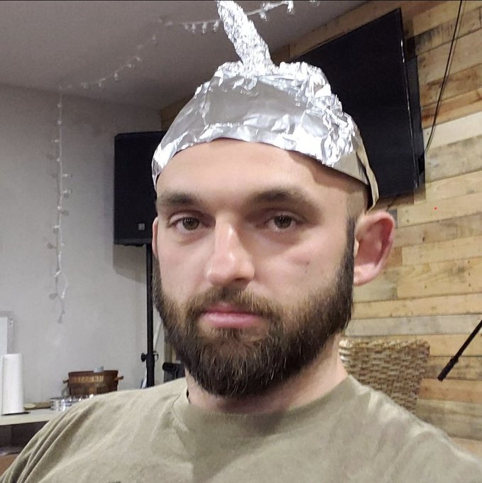 A bearded man wears a makeshift tinfoil hat with a crumpled peak on top, embodying the "tinfoil hat conspiracy" theme referenced in the post about blaming banks and fund managers for the CC exploit targeting Coinkite and NVK.