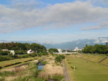 iced over river, green grassy field, white building with red roof, mountains in the distance, and a clear blue sky.