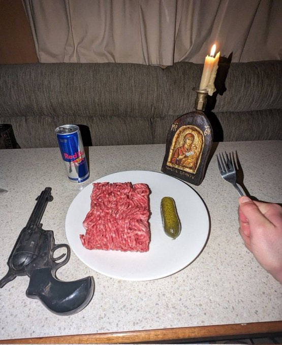 Surreal dining scene featuring a plate of raw ground meat and pickle next to a Red Bull can, a revolver, and a religious candle bottle with a religious icon, with a hand holding a fork.
