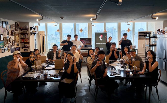 A group of about 15 people, many holding cookies, gather around a long table in a modern, high-rise loft with city views, posing for a group photo during the "Avalanche Bakery" event where they baked cookies while discussing RWA (Real World Asset) tokenization. The "Avalanche" logo is visible on a screen in the background, tying the casual baking gathering to the blockchain project's brand.