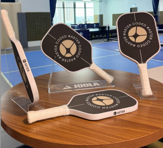 Four black and white pickleball paddles branded with 'ASTER CODED' are displayed on a wooden table at what appears to be a JOOLA facility.