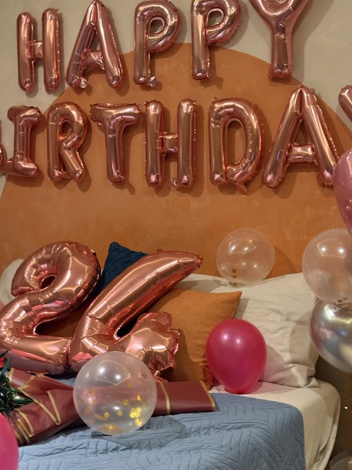 A bedroom celebration setup with rose-gold 'HAPPY BIRTHDAY' letter balloons, a large '24' balloon, and assorted pink and clear balloons scattered on the bed.