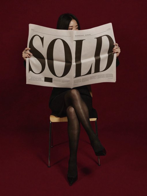A woman in black dress and stockings sitting on a stool against a dark red background, holding up a newspaper with 'SOLD' printed in large letters.