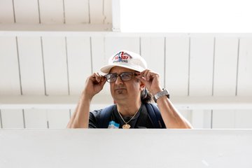 A man wearing a white cap and glasses is adjusting his eyewear in front of a white slatted background.