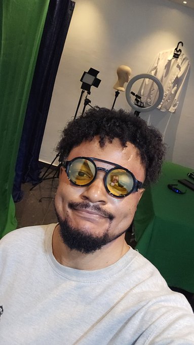 A selfie of a smiling man with curly hair and a beard wearing yellow-tinted round sunglasses, with studio lighting equipment visible in the background.