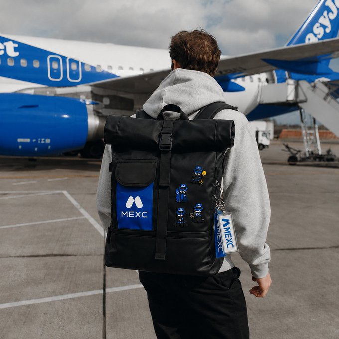 "The image features a man wearing a gray hoodie, carrying a backpack with a blue and white design that has the word 'excers' written on it. The background of the photo shows an airplane parked at an airport terminal."