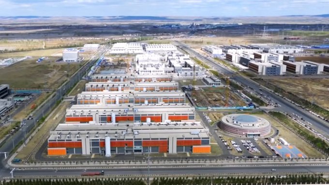 An aerial view of a large industrial complex or factory facility with multiple buildings featuring orange accents, situated in an expansive area with a distinctive circular building.