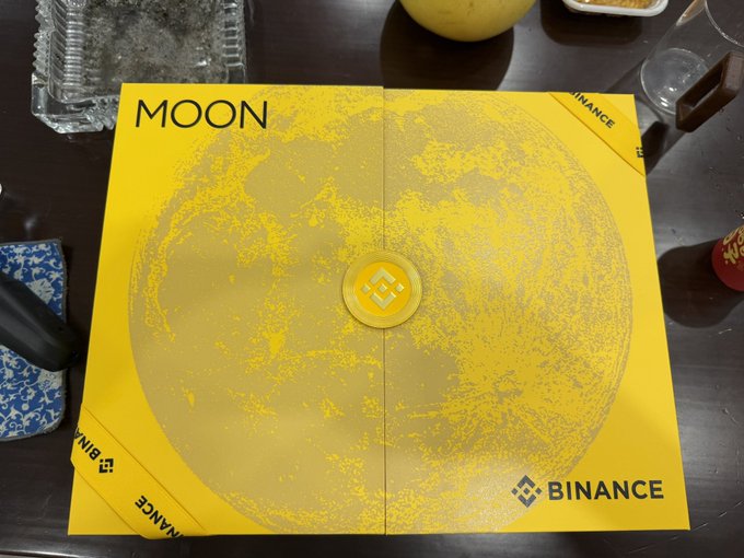 A yellow Binance-branded box labeled "MOON" featuring a moon design sits on a table alongside other items.