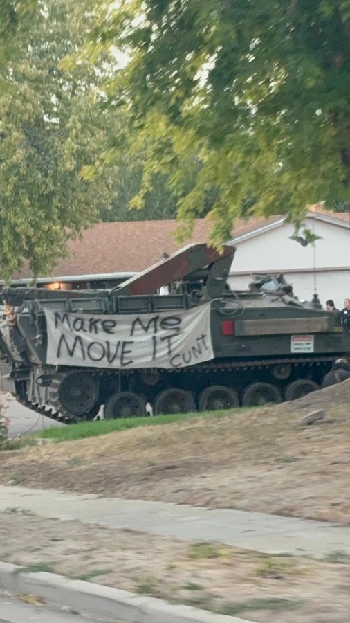An olive-green military tracked vehicle is parked outdoors with a hand-painted white banner hanging on its side that reads 'MAKE ME MOVE IT CUNT'.