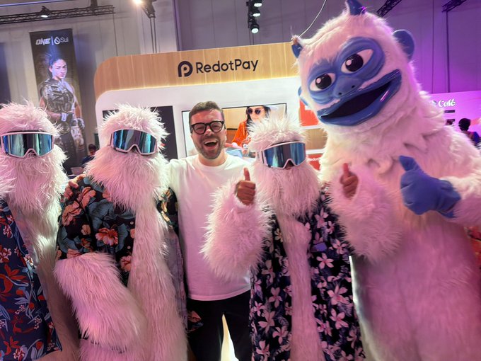 A man in a pink shirt poses with costumed mascots at a RedotPay booth during what appears to be a conference or expo event.