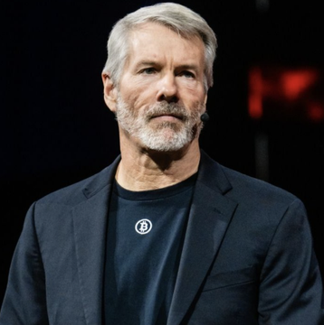 Portrait of a gray-haired man with a beard wearing a dark blazer over a black shirt with a Bitcoin logo, appearing to be at a speaking event.