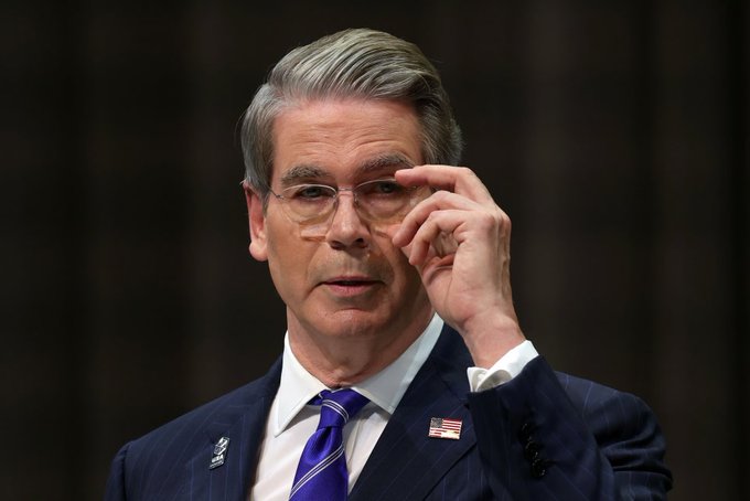 A man in a dark pinstripe suit and purple tie adjusts his glasses during what appears to be a formal hearing or testimony.