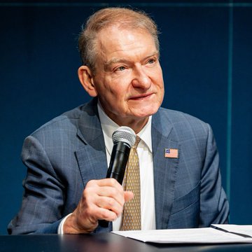 An older man with gray hair, wearing a blue checkered suit and an American flag lapel pin, holds a microphone while speaking at what appears to be a formal event. The image corresponds to the post about SEC Chair Paul Atkins advocating for the Senate to pass the CLARITY Act.