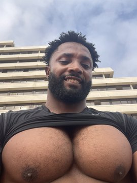 A selfie of a bearded man outdoors in front of a multi-story building with a cloudy sky behind him.