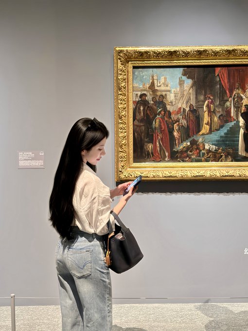 A young woman with long dark hair, wearing a cream blouse and denim skirt, stands in a museum or gallery looking at her phone in front of an ornately framed classical painting.