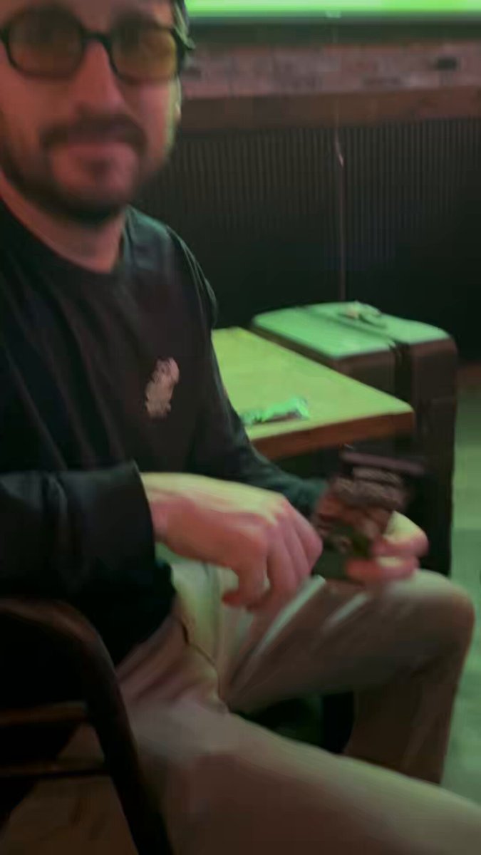 The image features a man sitting at a table, wearing glasses and holding a drink. The background shows a wooden wall with a green neon sign that reads "Rip some @MeebitsNFTs packs from @sergitosergito Watch the World Cup with some @BoredApeYC Drink some beers A Thursday evening in Brooklyn". The man is sitting on a chair, and there are two other chairs visible in the scene.