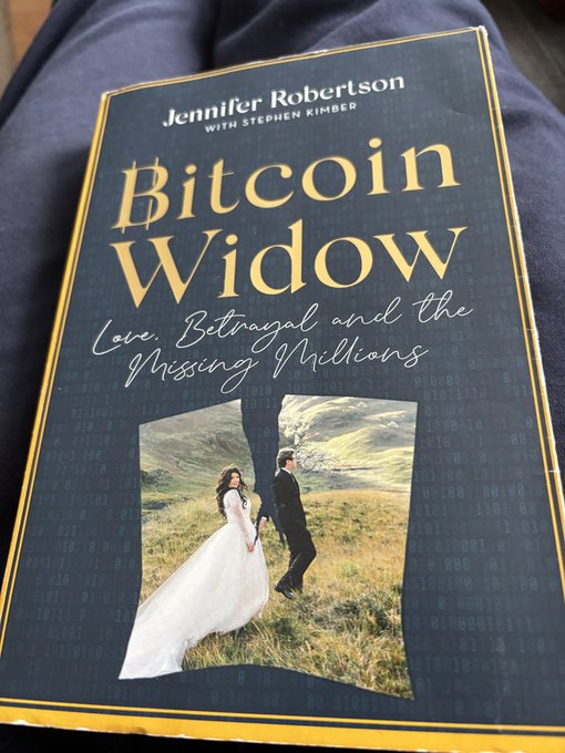 A book titled "Bitcoin Widow: Love, Betrayal, and the Missing Millions" by Jennifer Robertson with Stephen Kimber rests on dark fabric, its cover featuring a wedding photo of a couple framed by digital code patterns. The book tells the story of Robertson, whose husband Gerald Cotten, founder of the Canadian crypto exchange QuadrigaCX, died in India in 2018—taking the only passwords to the exchange's cold wallets with him.