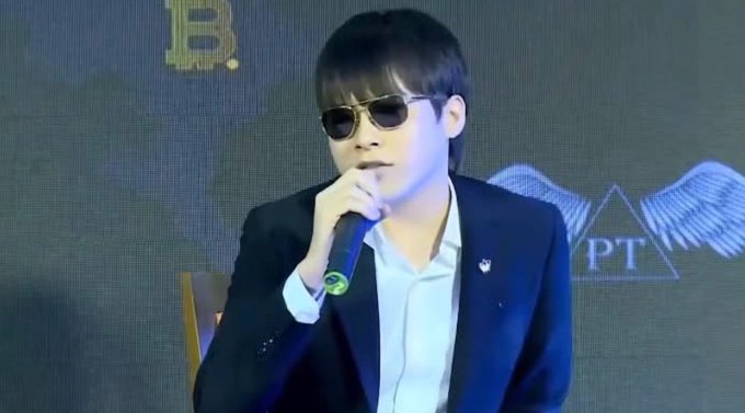 A man in a dark suit, white shirt, and sunglasses speaking into a microphone, with a 'BPT' winged logo visible on the screen behind him.